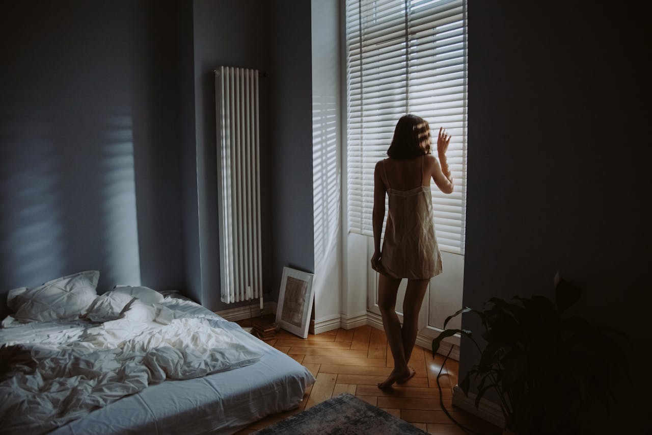 about-04 A woman in a bedroom stands by window blinds, casting shadows in soft morning light.